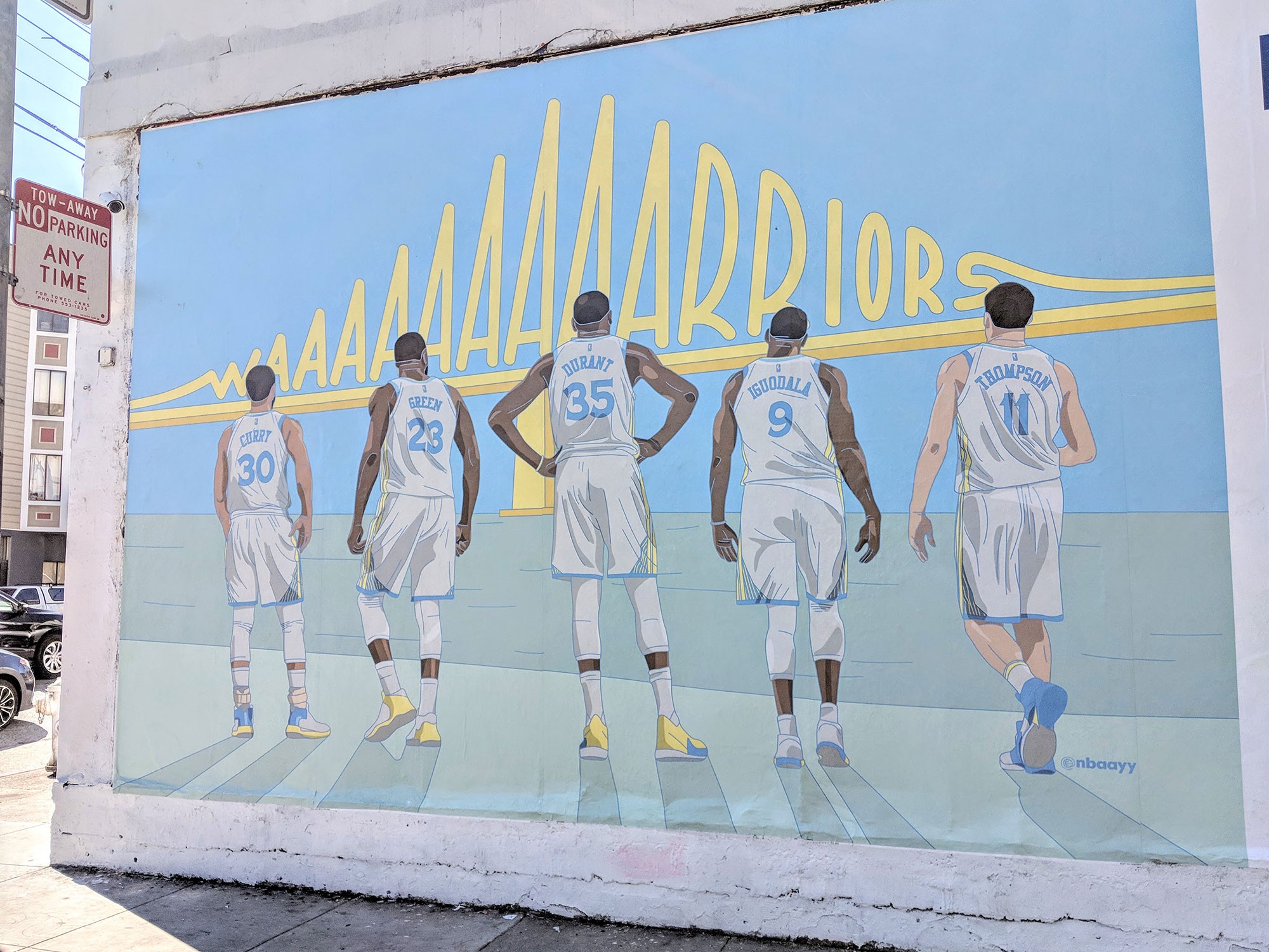 Street mural of five Warriors — Curry, Green, Durant, Iguodala and Thompson — facing a yellow Bay Bridge whose spans spell out Warriors.